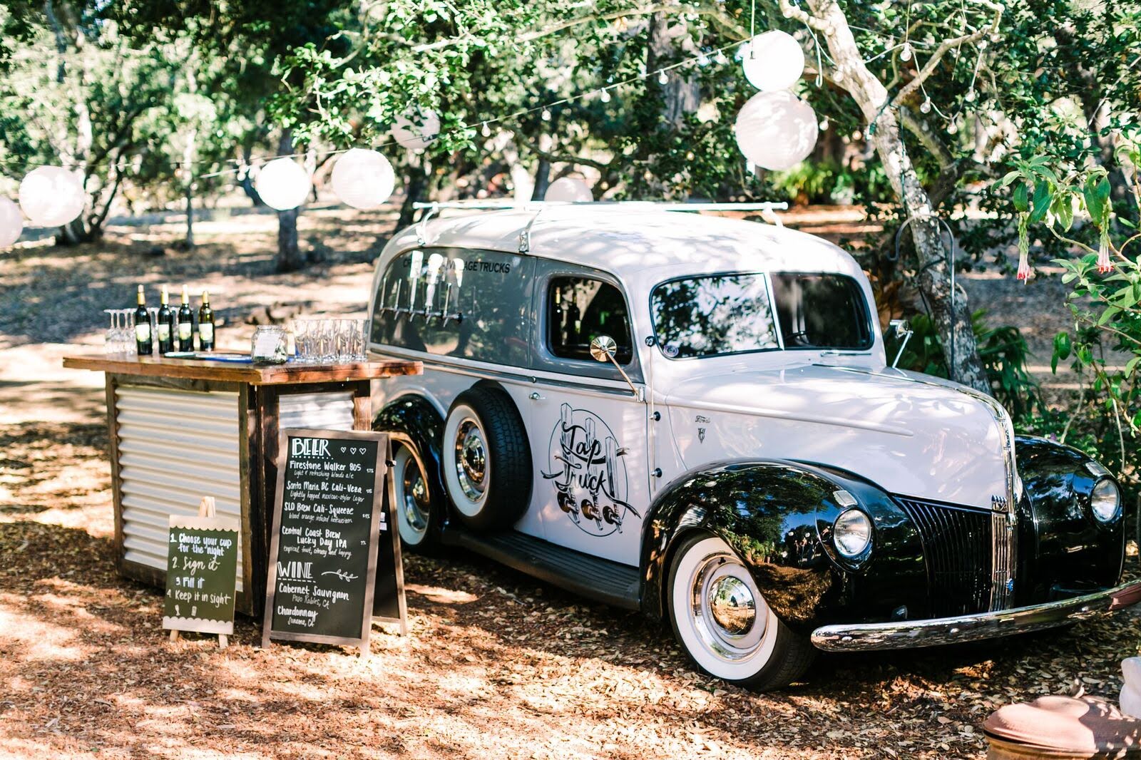 A vintage Tap Truck set up outdoors at an event, with taps on the side of the truck and a bar area nearby. The bar features bottles of wine, glasses, and a small chalkboard menu listing beverages. The scene is decorated with string lights and paper lanterns, creating a festive atmosphere in a wooded area.